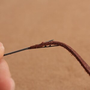 May include: A close-up of a brown leather cord being threaded with a needle. The needle is thin and sharp, and the leather is soft and pliable.