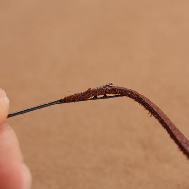 May include: A close-up of a brown leather cord being threaded with a needle. The needle is thin and sharp, and the leather is soft and pliable.