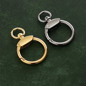 Puede incluir: Dos llaveros, uno dorado y otro plateado, con un anillo circular y un cierre giratorio. Los llaveros están sobre un fondo verde.