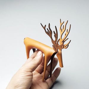 May include: A tan leather deer sculpture with large, intricately cut brown antlers. The deer is standing on a person's hand.