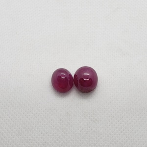 May include: Two polished, round, deep red gemstones. The gems have a smooth, reflective surface, and are set against a white background. The stones are similar in size and shape, and appear to be cabochons.