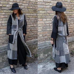 Può includere: Un lungo cappotto grigio e nero a patchwork con una cintura nera e un cappello nero. Il cappotto è realizzato con tessuti diversi e ha un aspetto unico e fatto a mano.
