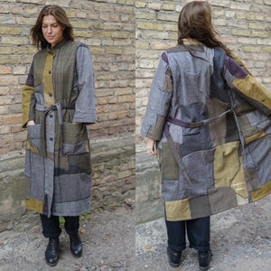 Può includere: Un lungo cappotto patchwork con vita a cintura. Il cappotto è realizzato con diversi tessuti in tonalità di marrone, grigio e verde. Il cappotto ha un colletto e maniche lunghe.