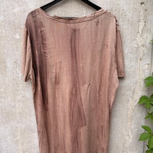 May include: A short-sleeved t-shirt in a muted, dusty brown color featuring a distressed, uneven wash pattern with darker brown vertical streaks and small, scattered holes throughout the fabric, displayed on a black plastic hanger.