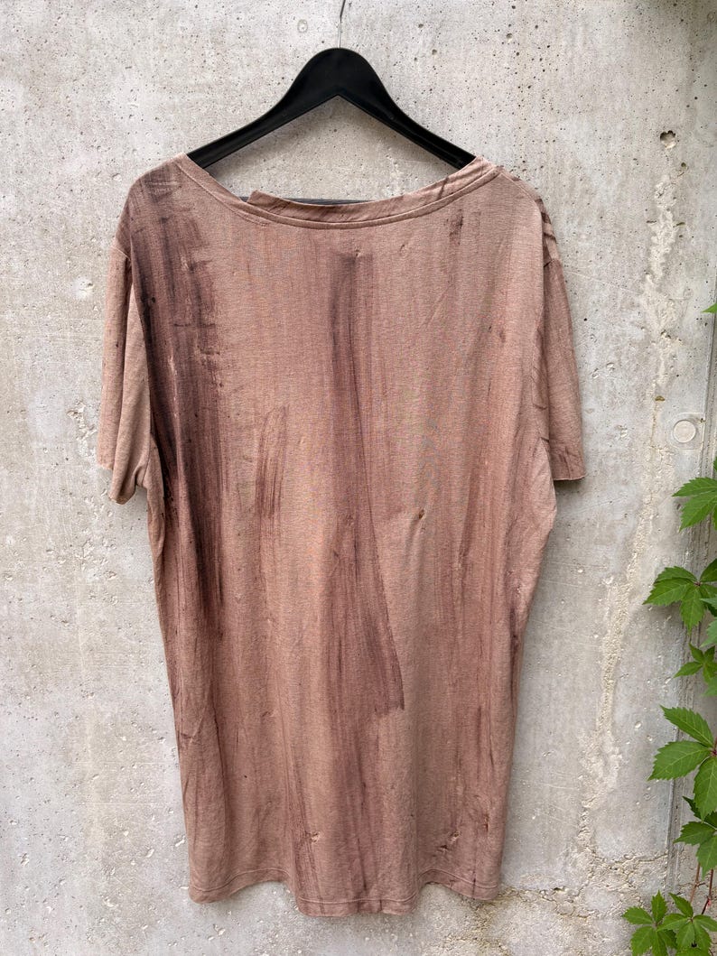 May include: A short-sleeved t-shirt in a muted, dusty brown color featuring a distressed, uneven wash pattern with darker brown vertical streaks and small, scattered holes throughout the fabric, displayed on a black plastic hanger.