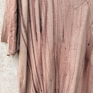 May include: A short-sleeved t-shirt in a light tan color featuring a dark brown vertical streaked pattern. The fabric has a textured appearance with visible dark brown splatters and vertical lines running down the front of the garment.