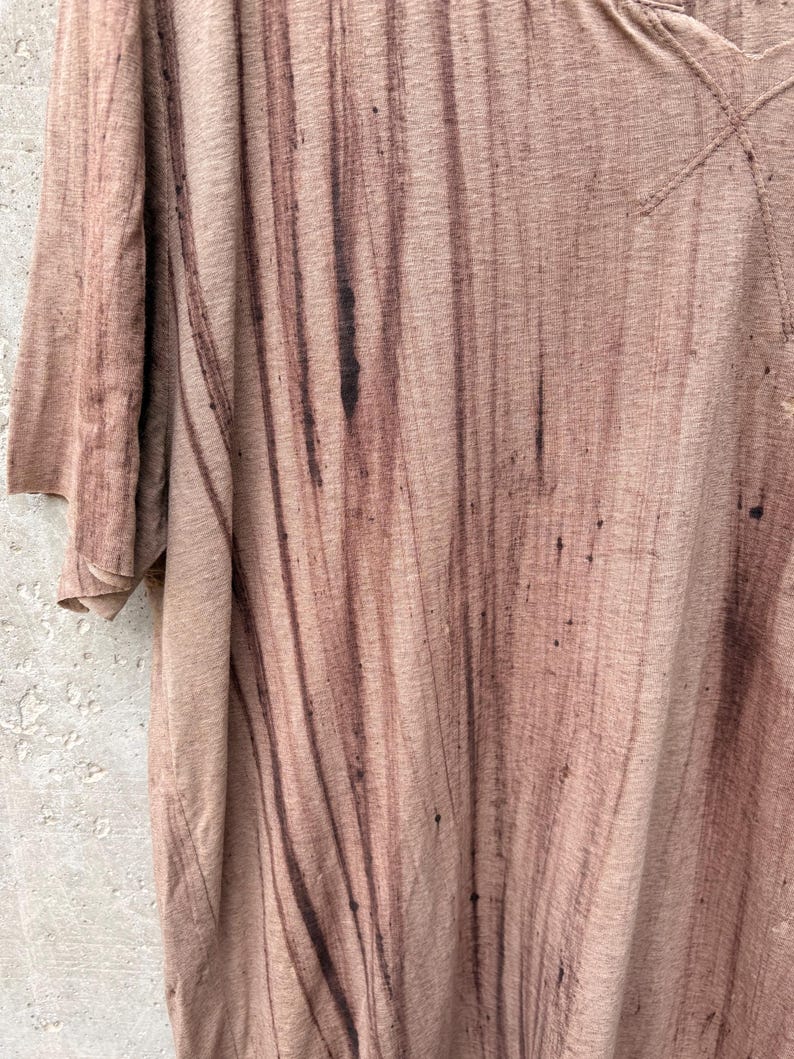 May include: A short-sleeved t-shirt in a light tan color featuring a dark brown vertical streaked pattern. The fabric has a textured appearance with visible dark brown splatters and vertical lines running down the front of the garment.