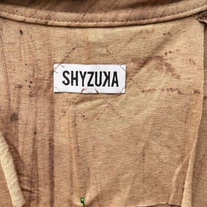 May include: A tan-colored garment featuring a white rectangular fabric label stitched onto the center with the black text SHYZUXA. The fabric has a mottled, uneven brown dye pattern and raw, unfinished edges around the neck and label area.