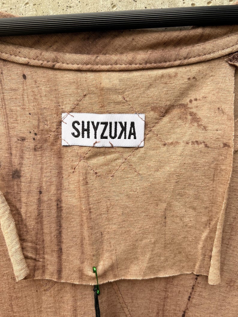 May include: A tan-colored garment featuring a white rectangular fabric label stitched onto the center with the black text SHYZUXA. The fabric has a mottled, uneven brown dye pattern and raw, unfinished edges around the neck and label area.