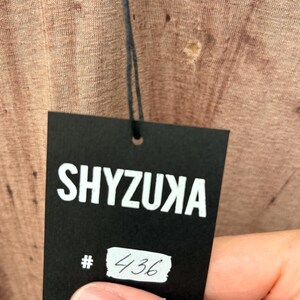 May include: A textured light brown fabric with a visible weave pattern attached to a black rectangular paper tag. The tag features the white text SHYZUXA printed at the top and a white rectangular sticker with the handwritten number 436 below it.