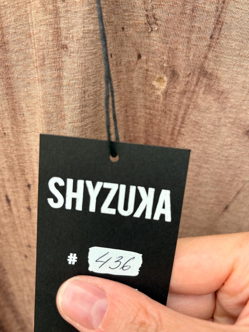 May include: A textured light brown fabric with a visible weave pattern attached to a black rectangular paper tag. The tag features the white text SHYZUXA printed at the top and a white rectangular sticker with the handwritten number 436 below it.
