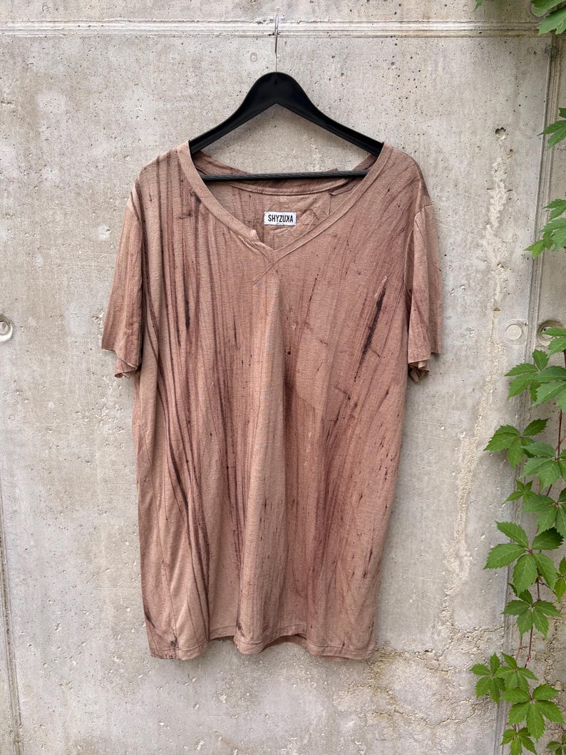 May include: A short-sleeved V-neck t-shirt in a muted brown color with a vertical black streaked pattern. The garment features a white interior neck label with the black text SHYZUXA and is displayed on a black plastic hanger.