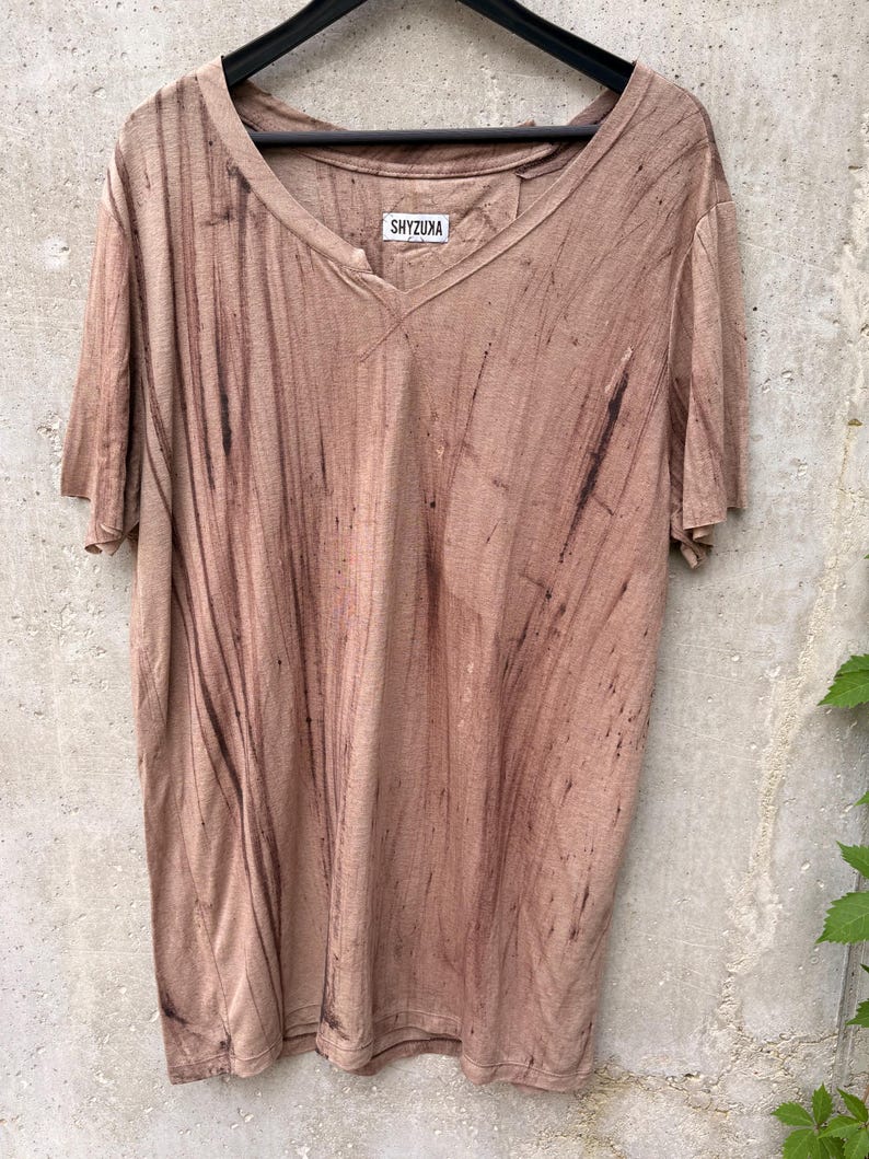 May include: A short-sleeved V-neck t-shirt in a muted tan color with dark brown vertical streaks throughout the fabric. The shirt features a white label at the inner neckline with the black text SHYZUXA printed on it, hanging on a black hanger.