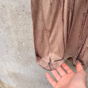 May include: A light brown t-shirt featuring a vertical dark brown distressed or paint-splatter effect pattern. The garment has a visible side seam and a hemmed bottom edge, showcasing a textured fabric appearance with a subtle, uneven color distribution.