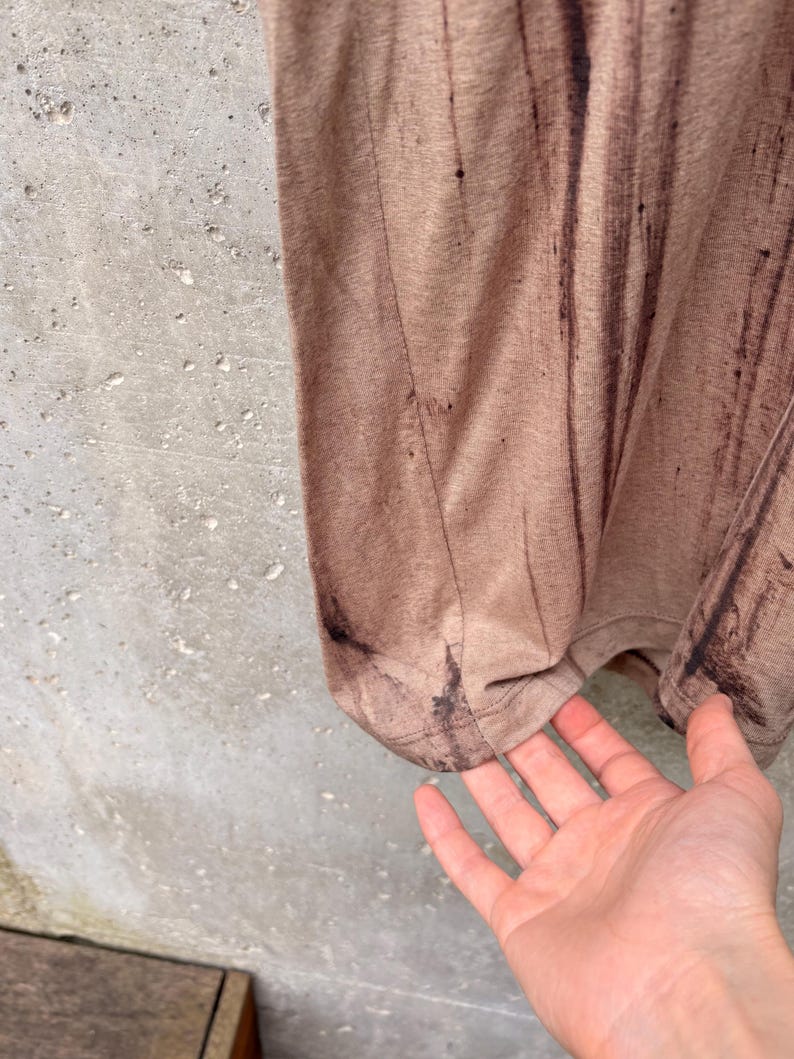 May include: A light brown t-shirt featuring a vertical dark brown distressed or paint-splatter effect pattern. The garment has a visible side seam and a hemmed bottom edge, showcasing a textured fabric appearance with a subtle, uneven color distribution.