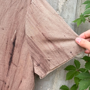 May include: A short-sleeved t-shirt in a light brown or tan color featuring a dark brown, streaky, wood-grain-like pattern throughout the fabric. The garment has a simple, straight-cut sleeve and a lightweight, textured appearance.