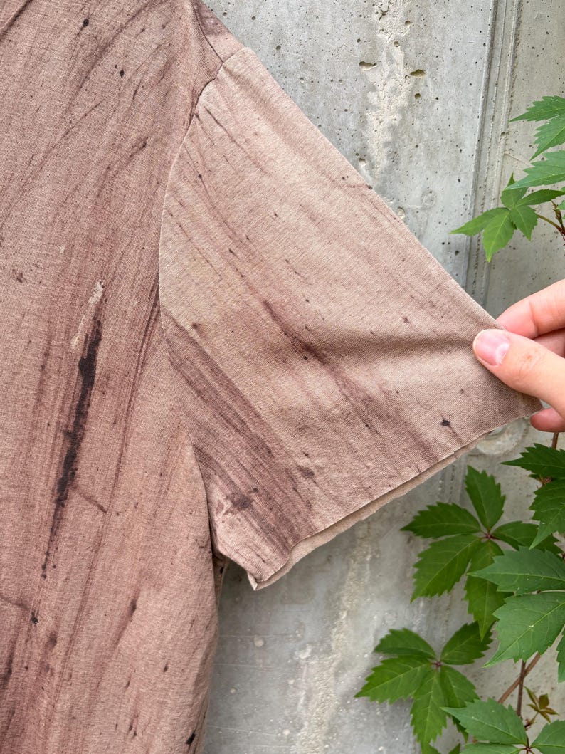 May include: A short-sleeved t-shirt in a light brown or tan color featuring a dark brown, streaky, wood-grain-like pattern throughout the fabric. The garment has a simple, straight-cut sleeve and a lightweight, textured appearance.