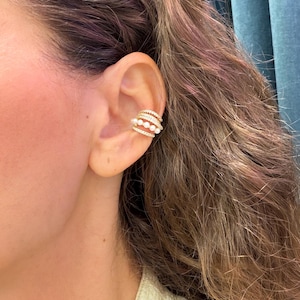 May include: A gold ear cuff featuring multiple bands. The cuff is adorned with small, white pearls and clear, sparkling stones. The jewelry is worn on the ear, with the person's hair visible in the background.