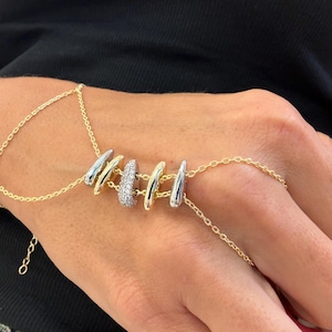 May include: A gold hand chain with a series of silver and gold-toned curved bars. One bar is encrusted with small, clear stones. The chain connects to a ring and a bracelet, creating a unique jewelry piece.