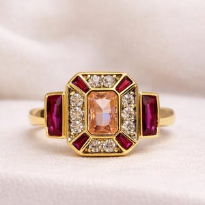 May include: A gold-toned ring with a central peach-colored rectangular gemstone, surrounded by smaller white and red gemstones. Two rectangular red gemstones flank the central design. A classic, elegant jewelry piece.