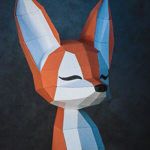 Peut inclure: Sculpture g&eacute;om&eacute;trique en papier repr&eacute;sentant un renard, avec des sections orange, blanches et bleues. Le renard a les yeux ferm&eacute;s et un nez noir. L'&oelig;uvre est pos&eacute;e sur un fond sombre.