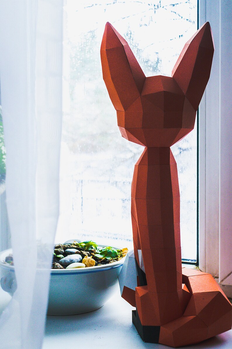 Peut inclure: Une sculpture g&eacute;om&eacute;trique en papier repr&eacute;sentant un renard, dans des tons orange et marron, est pos&eacute;e sur un rebord de fen&ecirc;tre. Le renard est &agrave; c&ocirc;t&eacute; d'un petit bol avec des pierres et de la verdure.