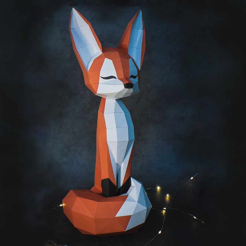 Peut inclure: Une sculpture de renard en papier aux tons orange, blanc et gris, les yeux ferm&eacute;s, assis droit. Le renard a un design g&eacute;om&eacute;trique et polygonal. Le fond est une surface sombre et textur&eacute;e, avec des guirlandes lumineuses.