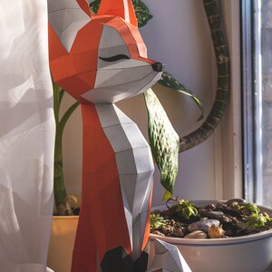 Peut inclure: Une sculpture de renard g&eacute;om&eacute;trique en papier, aux tons orange, blanc et gris. Le renard a les yeux ferm&eacute;s et se trouve pr&egrave;s d'une fen&ecirc;tre avec des plantes. C'est un objet d&eacute;coratif pour la maison ou le bureau.