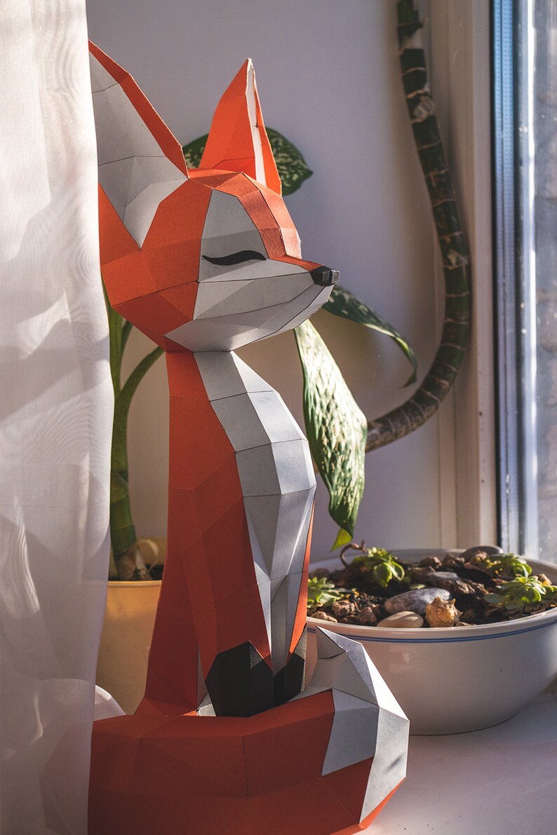 Peut inclure: Une sculpture de renard g&eacute;om&eacute;trique en papier, aux tons orange, blanc et gris. Le renard a les yeux ferm&eacute;s et se trouve pr&egrave;s d'une fen&ecirc;tre avec des plantes. C'est un objet d&eacute;coratif pour la maison ou le bureau.