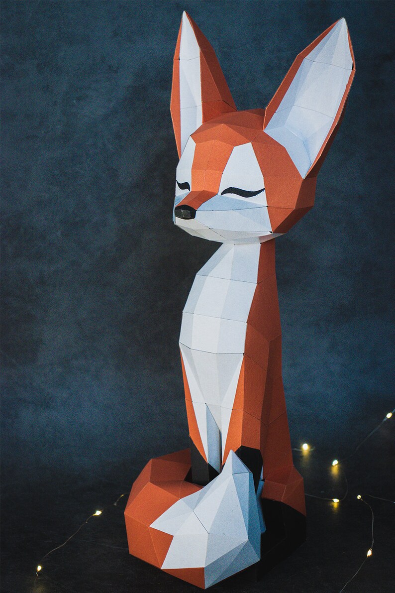 Peut inclure: Sculpture g&eacute;om&eacute;trique en papier repr&eacute;sentant un renard aux couleurs orange, blanc et noir. Le renard a un corps long et des oreilles pointues. Le renard est assis, les yeux ferm&eacute;s, ce qui lui donne une apparence paisible. Le fond est bleu fonc&eacute;.