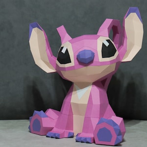 May include: A pink papercraft figure of a cartoon character with large ears, sitting with its legs crossed. The figure has black eyes, a purple nose, and purple accents on its feet. The figure is made of geometric shapes.