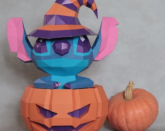 May include: A papercraft Stitch figure wearing a witch hat, emerging from an orange jack-o'-lantern. The figure is blue with pink ears and purple accents. A whole orange pumpkin sits beside the jack-o'-lantern, creating a Halloween theme.