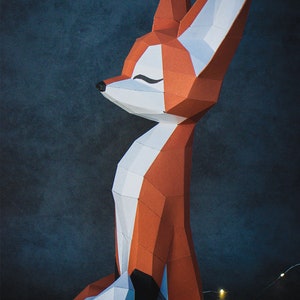 Peut inclure: Sculpture de renard g&eacute;om&eacute;trique en papier, aux tons orange, blanc et gris, sur fond bleu fonc&eacute;. Le renard a un design polygonal stylis&eacute;, les yeux ferm&eacute;s, et est en position assise. La sculpture est &eacute;clair&eacute;e par de petites guirlandes lumineuses.