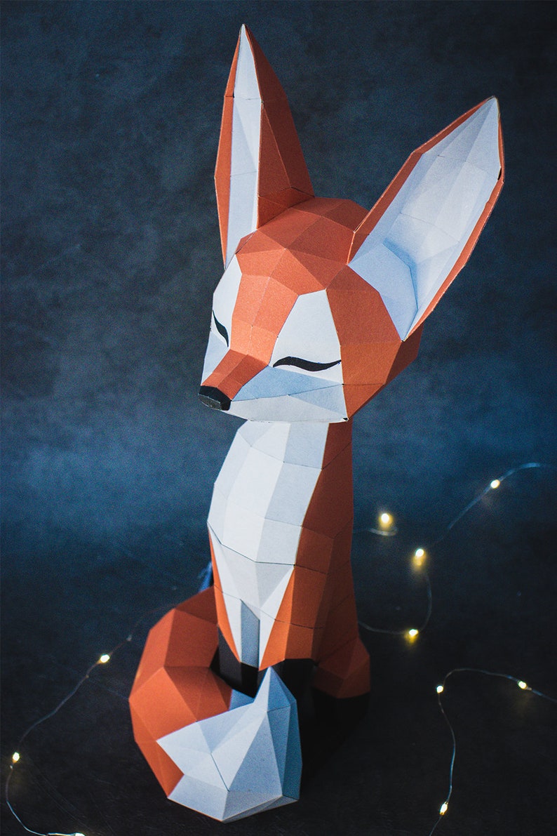 Peut inclure: Une sculpture g&eacute;om&eacute;trique en papier repr&eacute;sentant un renard, dans des tons d'orange, de blanc et de gris. Le renard est assis, les yeux ferm&eacute;s, avec un design moderne et polygonal. Le fond est bleu-gris fonc&eacute;, avec quelques petites lumi&egrave;res.
