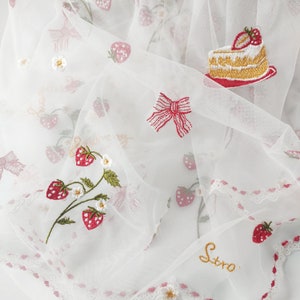 Puede incluir: Tela blanca transparente con bordados de fresas, lazos y una porci&oacute;n de pastel. La tela presenta un delicado ribete de encaje y la palabra "Stro" bordada con hilo dorado. El dise&ntilde;o incluye acentos rojos, verdes y blancos.