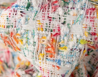 Puede incluir: Primer plano de una tela blanca con un patrón tejido de colores. La tela tiene un aspecto texturizado y peludo y presenta una variedad de colores, incluyendo rojo, naranja, amarillo, verde, azul y morado.
