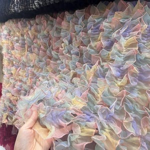 May include: A close-up of a pastel-colored fabric with ruffled layers. The fabric features a mix of soft colors, including pink, yellow, and lavender. The ruffled texture creates a delicate and airy appearance, suitable for clothing or decorative projects.
