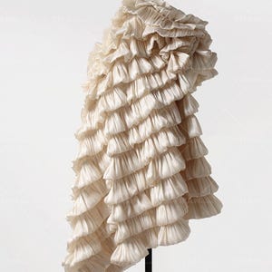 Puede incluir: Una prenda de color crema con volantes y un diseño en cascada de capas. La prenda está hecha de una tela suave y fluida y tiene una forma asimétrica única.