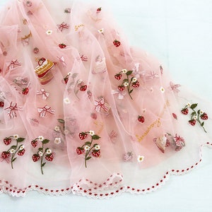 Puede incluir: Tela rosa p&aacute;lido transparente decorada con bordados de fresas, lazos y motivos de pastel. La tela presenta un borde festoneado con detalles rojos y blancos. La palabra "strawberry" est&aacute; escrita en cursiva dorada.