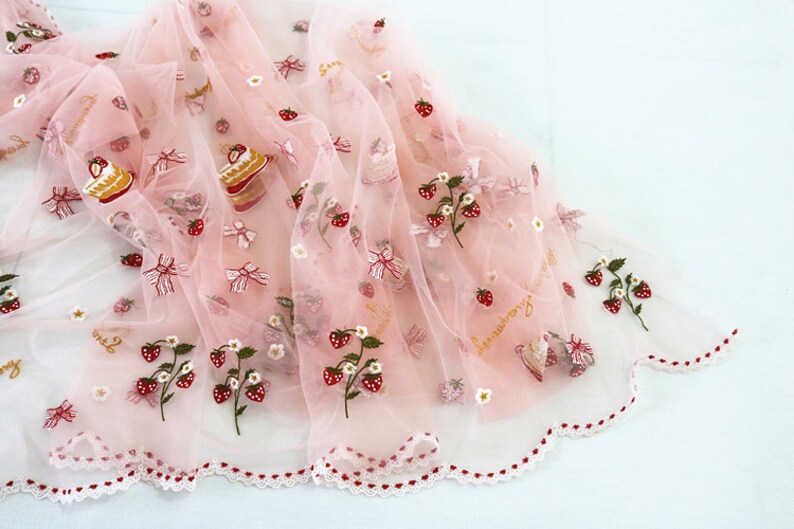 Puede incluir: Tela rosa p&aacute;lido transparente decorada con bordados de fresas, lazos y motivos de pastel. La tela presenta un borde festoneado con detalles rojos y blancos. La palabra "strawberry" est&aacute; escrita en cursiva dorada.