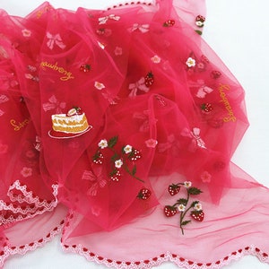 Puede incluir: Una tela transparente de color rosa intenso con bordados de fresas, lazos y dise&ntilde;os de pastel. La palabra "strawberry" est&aacute; bordada en hilo dorado. Los bordes est&aacute;n adornados con un borde festoneado.