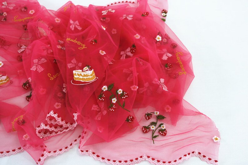Puede incluir: Una tela transparente de color rosa intenso con bordados de fresas, lazos y dise&ntilde;os de pastel. La palabra "strawberry" est&aacute; bordada en hilo dorado. Los bordes est&aacute;n adornados con un borde festoneado.