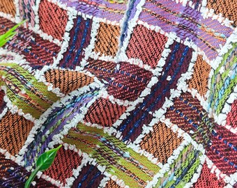 Puede incluir: Primer plano de una tela tejida colorida con un patrón a cuadros. La tela está hecha de hilos de diferentes colores, incluyendo rojo, verde, morado y blanco. La tela tiene una superficie texturizada y un borde con flecos.