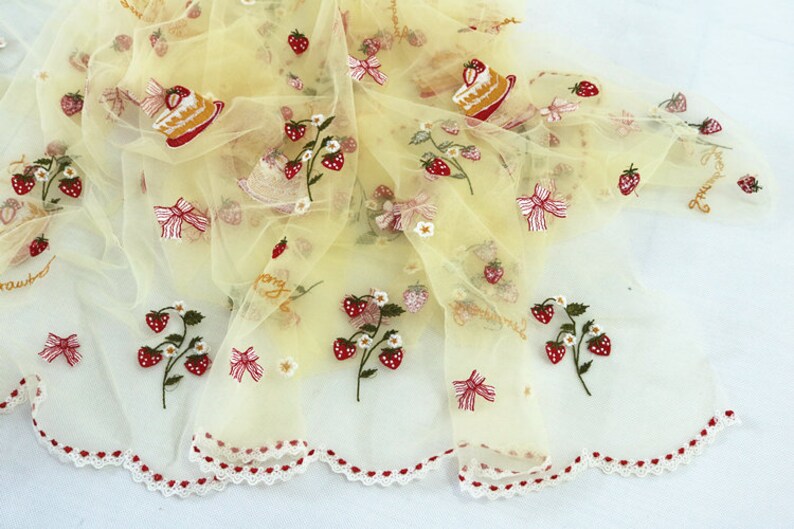 Puede incluir: Una tela transparente de color amarillo p&aacute;lido con bordados de fresas, lazos y rebanadas de pastel. La tela tiene un borde festoneado con detalles rojos en forma de coraz&oacute;n. La palabra "strawberry" est&aacute; bordada en cursiva.