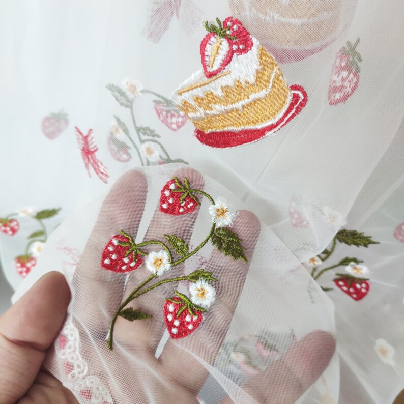 Puede incluir: Tela blanca transparente con bordados de fresas, flores y un dise&ntilde;o de pastel. El bordado utiliza hilos rojos, verdes, blancos y amarillos. La tela se sostiene en una mano, mostrando los delicados detalles del dise&ntilde;o.