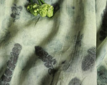 Puede incluir: Primer plano de una tela verde claro con estampados botánicos en gris oscuro. La tela está drapeada, con pliegues y texturas visibles. Un pequeño racimo de flores verdes descansa sobre la tela, añadiendo un elemento natural.