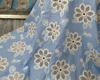 May include: Light blue fabric with a repeating pattern of white, openwork floral designs. The flowers are detailed with a central pattern and surrounded by leaves and smaller floral accents. The fabric is gathered, creating soft folds.