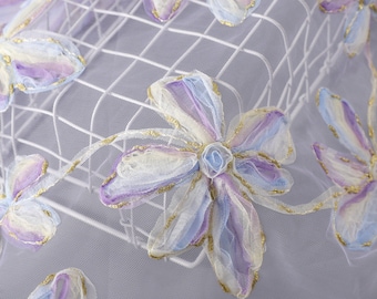 Può includere: Primo piano di un tessuto delicato e iridescente con fiori ricamati in tonalità di viola, blu e bianco. I fiori hanno un leggero luccichio dorato.