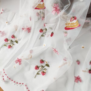 Puede incluir: Tela blanca transparente bordada con fresas rojas, lazos y rebanadas de pastel. El bordado incluye la palabra "strawberry" en escritura dorada. La tela tiene un borde festoneado con ribete rojo y peque&ntilde;as flores blancas.