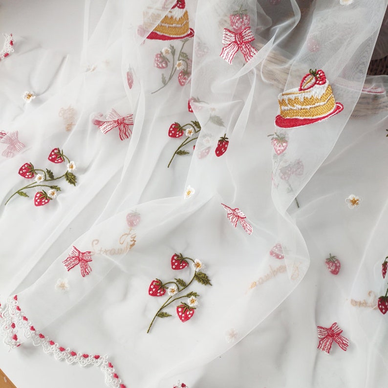 Puede incluir: Tela blanca transparente bordada con fresas rojas, lazos y rebanadas de pastel. El bordado incluye la palabra "strawberry" en escritura dorada. La tela tiene un borde festoneado con ribete rojo y peque&ntilde;as flores blancas.